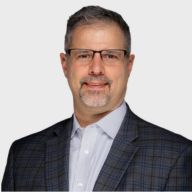 A middle-aged man with short gray hair, glasses, and a goatee, wearing a checked suit jacket and light-colored collared shirt, posing against a plain white background. EFESO Management Consultants